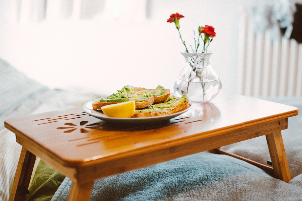 A cozy breakfast setting featuring avocado toast on a wooden tray with flowers in a vase.
